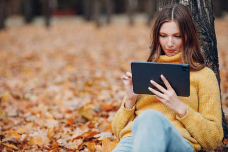 Young woman model sitting with tablet pc in autumn park with yellow foliage maple leaves. Fall season fashionの写真素材