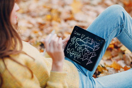 Young woman model sitting with tablet pc and painting with pencil stylus in autumn park with yellow foliage maple leaves.の写真素材