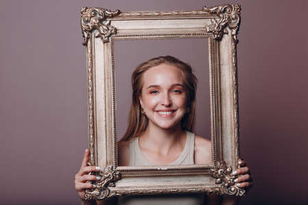 Millenial young woman blonde hair holds gilded picture frame in hands face portrait.の写真素材