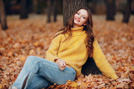 Young woman model sitting in autumn park with yellow foliage maple leaves. Fall season fashionの写真素材