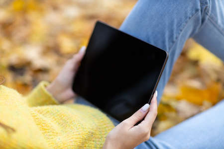 Young woman model buying online shopping with digital tablet pad in autumn park with yellow foliage maple leaves. Fall season fashion mockup.の写真素材