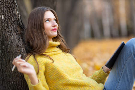 Young woman model sitting with tablet pc and painting with pencil stylus in autumn park with yellow foliage maple leaves.の写真素材