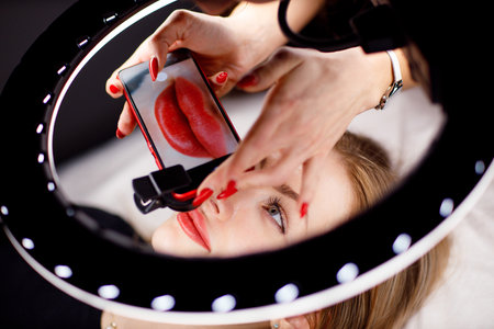Cosmetologist making photo of work on mobile phone red permanent make up tattoo on young woman lips.の写真素材