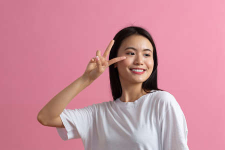 Portrait of asian positive smiling young woman with V fingers gesture on pink background.の写真素材