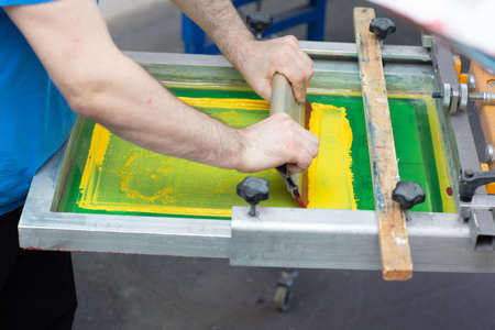 Serigraphy silk screen print process at clothes factory. Frame, squeegee and plastisol color yellow paintsの写真素材