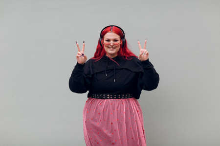 European plus size woman in headphones showing V victory fingers sign. Young red pink haired body positive teenager.の写真素材