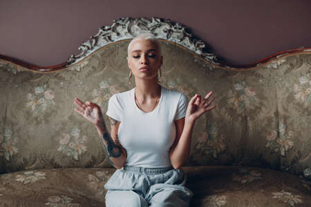 Millenial young woman with short blonde hair portrait sitting and meditate with ok sign mudra on vintage sofaの写真素材