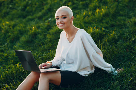 Portrait of young smiling millenial european short haired woman using laptop at green grass meadow in park. Beautiful happy blonde girl outdoor. Summer fashion female clothing.の写真素材