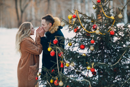 Young adult couple decorates christmas tree in winter forest and warms hands with breath. New year pine holiday party celebrationの写真素材