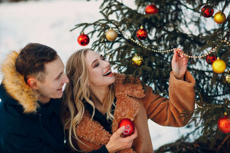 Young adult couple decorates christmas tree in winter park wood fir forest. New year pine holiday party celebration concept.の写真素材