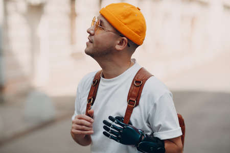 Young disabled man with artificial prosthesis hand in casual clothes and backpack walking at sunny city street outdoorの写真素材