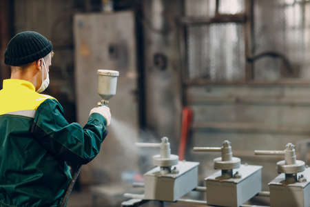 Powder primer coating of metal parts. Worker man in a protective suit sprays powder paint from gun on metal product construction at factory plant.の写真素材