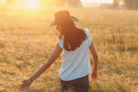 Woman farmer in cowboy hat walking with hands on ears at agricultural rye field on sunset.の写真素材