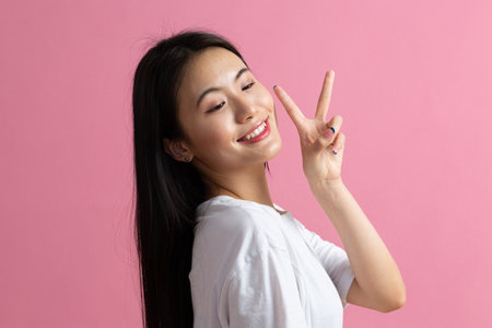 Portrait of asian positive smiling woman with V fingers gesture on pink background.の写真素材