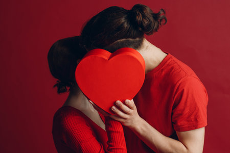 Man and woman young couple kissing and hiding behind heart-shaped box.の写真素材