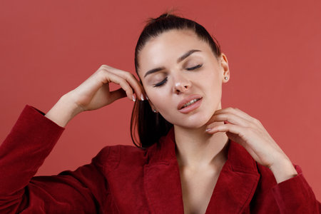 Young smiling caucasian 30s woman doing face skin yoga massage in red velvet jacket portrait.の写真素材