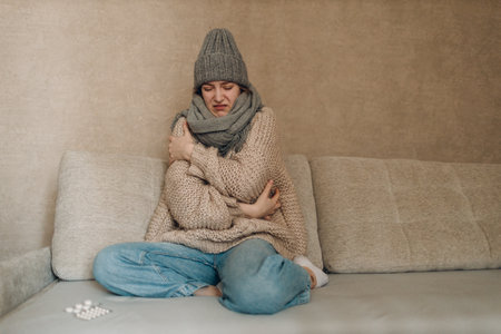 Young woman wearing scarf and wool hat has flu ill sick disease cold at home indoor on sofaの写真素材