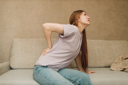 Young woman with back spine pain ache sitting on sofaの写真素材