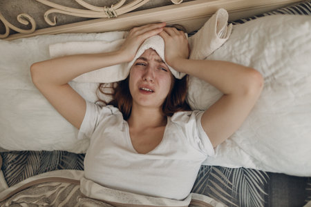 Young woman with headache flu ill sick disease cold at home indoor lying on bed with cold compress on her head at bedroom homeの写真素材