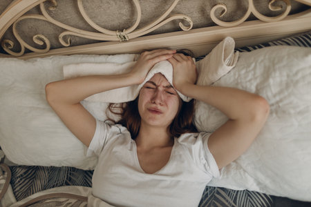 Young woman with headache flu ill sick disease cold at home indoor lying on bed with cold compress on her head at bedroom homeの写真素材