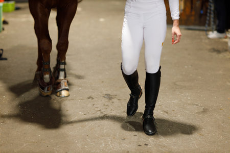 Female legs in black leather boots close up rider jockey walking with horse at stable and preparing horse racing or jumping competitionの写真素材