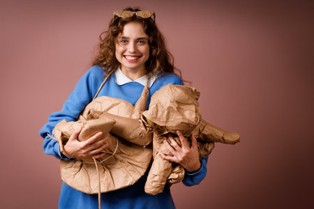 Young positive smiling woman receives surprise gift and holding in hands bunch of gifts wrapped in rustic beige craft paper. Hat, shoes, hair dryer, soft toy and glassesの写真素材