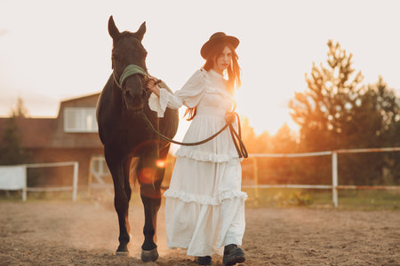 Woman in white dress and black hat with her horse at sunset outdoors ranchの写真素材