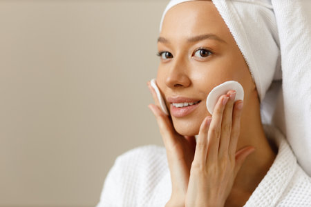 Young woman in white robe and towel on her head wipes the skin of her face with cotton pads. Cleansing and healthy body care after shower. Close-up of pretty girl on isolated background, copy space.の写真素材