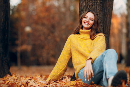 Beautiful girl at autumn park sitting near a tree with falling yellow foliage leaves. Woman smile.の写真素材