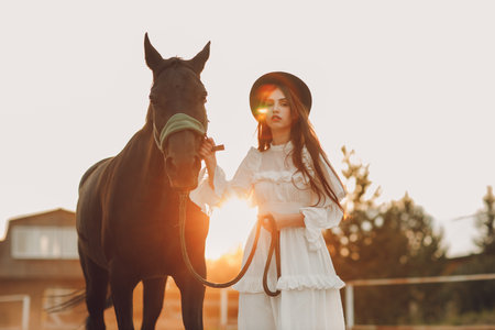Woman in white dress and black hat with her horse at sunset outdoors ranchの写真素材