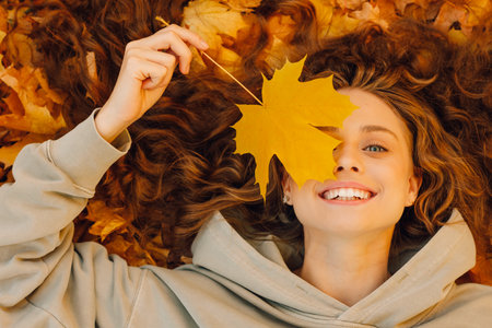 Smiling young woman lies on the autumn foliage in the park with the yellow leaves