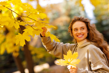 Beautiful smiling young woman enjoys the autumn weather at the maple tree with the yellow leaves at sunsetの写真素材