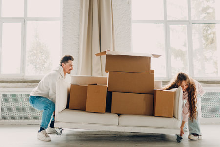 Happy young family couple man and woman push sofa with cardboard boxes in new home. Moving delivery concept.の写真素材