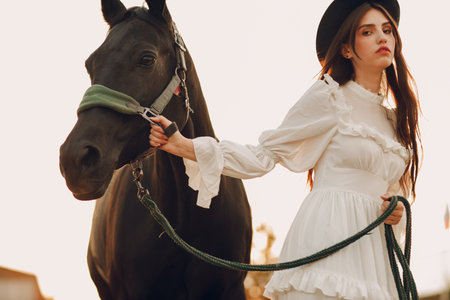 Woman in white dress and black hat with her horse at outdoors ranchの写真素材