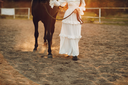 Woman in white dress and black hat with her horse at sunset outdoors ranchの写真素材