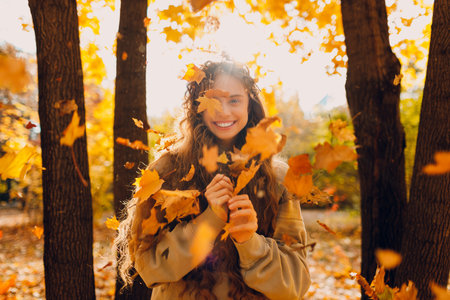 Smiling positive young woman looking to camera and throws up yellow autumn leaves in the forest at sunsetの写真素材