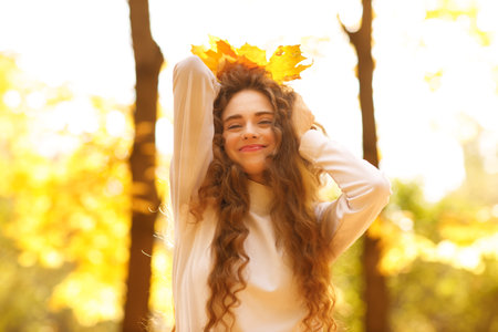 Smiling young woman with the fall leaves crown on her curly hairstyle head in the autumn park at sunsetの写真素材