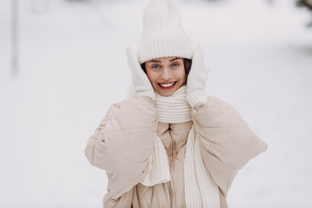 Happy smiling young woman put on scarf hat and mittens enjoys winter weather at snowy winter parkの写真素材