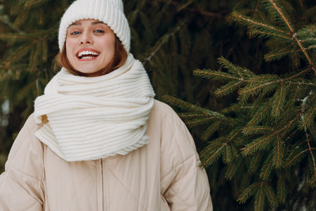 Happy smiling young woman portrait dressed coat scarf hat and mittens enjoys winter weather at winter park with pinesの写真素材