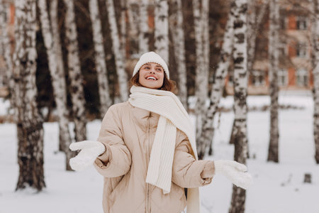 Young smiling woman dressed coat scarf hat and mittens enjoys winter weather at birch winter parkの写真素材