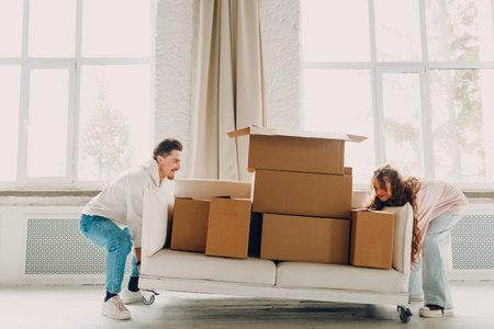 Happy young family couple man and woman push sofa with cardboard boxes in new home. Moving delivery concept.の写真素材