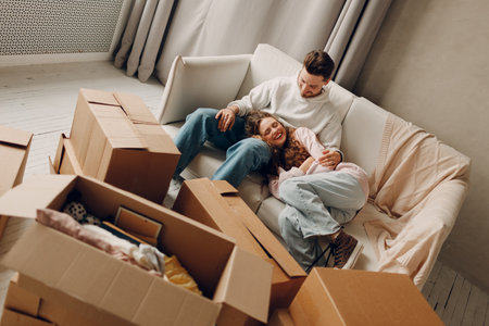 Happy young family couple man and woman relaxing on sofa after moving cardboard boxes to new estate...