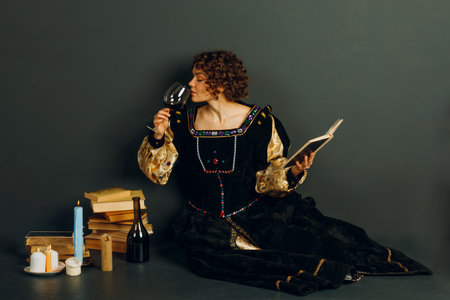 Young adult woman dressed in a medieval dress sitting near ancient books and candles with bottle of red wine and drinking a glass of wineの写真素材
