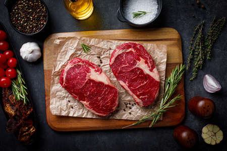 Raw pork steak pieces with spices and herbs rosemary, thyme, basil, salt and pepper on rustic wooden cutting board. Flat lay over dark black stone backgroundの写真素材