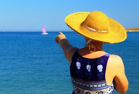 Gentle young woman on the sea, showing toward small sailing ship の写真素材