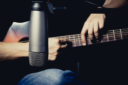 male musician playing acoustic guitar behind condenser microphone in recording studioの写真素材