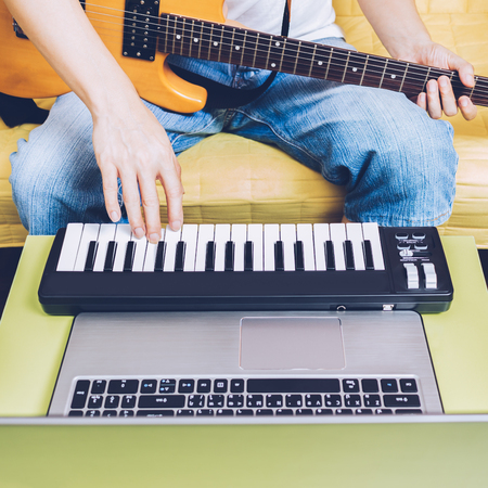male musician composing a song on computer laptop, midi keyboard and electric guitar in living room for trendy music production technology in modern lifestyleの写真素材