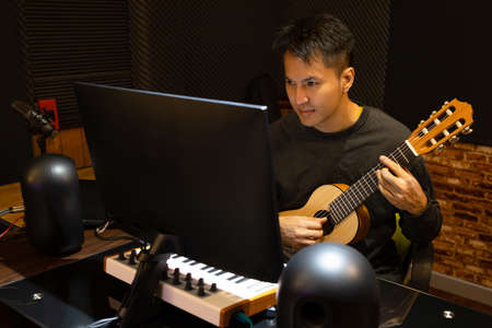 asian handsome male music vlogger streaming a live video while playing acoustic guitar at home or...