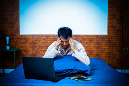 headache, tense young asian man working on laptop computer in bedroom at nightの写真素材