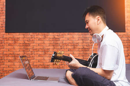 asian young man enjoy learning guitar lesson from internet on laptop computer in bedroomの写真素材